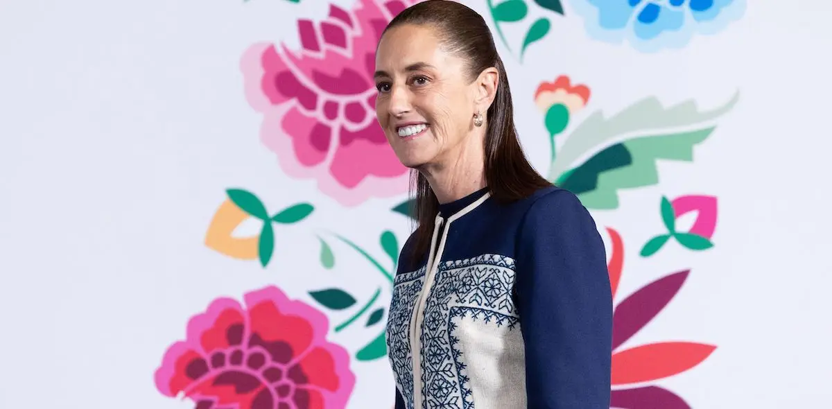 La presidenta Claudia Sheinbaum sonríe a los asistentes de la conferencia mañanera en Palacio Nacional. Hay un fondo blanco con rosas pintadas. 