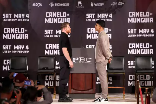 Foto de Canelo Álvarez frente a Jaime Munguía en la conferencia de prensa previa a la pelea que se disputará en Las Vegas. Álvarez viste un conjunto completamente negro con botas, mientras que Munguía lleva un conjunto color camel, con tenis y playera blanca.