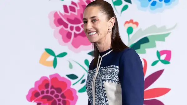 La presidenta Claudia Sheinbaum sonríe a los asistentes de la conferencia mañanera en Palacio Nacional. Hay un fondo blanco con rosas pintadas. 