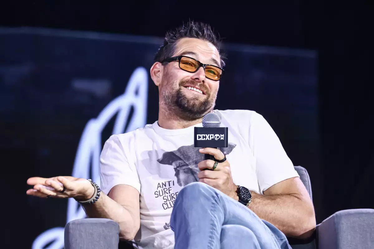 foto del actor Antony Starr con lentes amarillos y playera blanca, sosteniendo un micrófono trianguilar del CCXP México 2024.