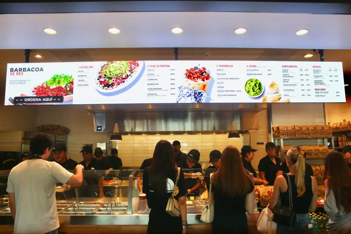 Mostrador de Chipotle con una fila de clientes que esperan para ordenar sus alimentos.
Al fondo, varios empleados preparan los pedidos detrás de una barra de servicio.
Sobre el mostrador se observa un menú con opciones de bowls, burritos, tacos y ensaladas.
A un costado hay estantes con bolsas de chips y otros complementos.