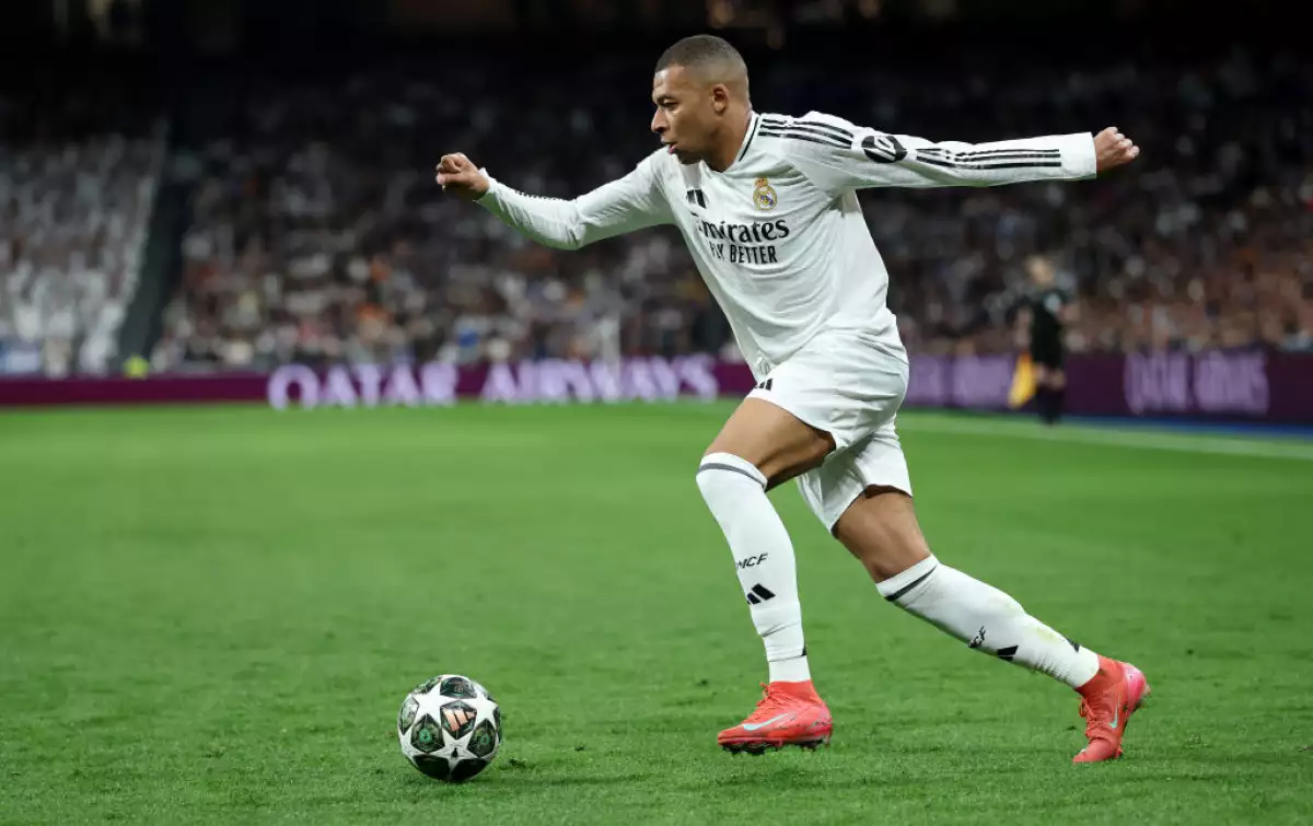 Foto de Kylian Mbappé en el campo de juego a punto de patear el balón de futbol, viste botines rojos y el uniforme del Real Madrid.