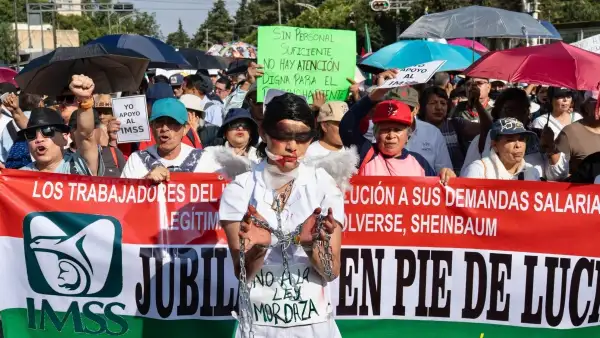 Trabajadores del IMSS protestando en una calle, varios sostienen una manta larga en la que exigen solución a sus demandas salariales; enfrente una persona con el uniforme blanco, las manos amarradas y la boca y los ojos tapados. 