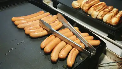 Una plancha industrial muestra varias salchichas dorándose, con pinzas metálicas colocadas sobre ellas. Al fondo, una segunda superficie caliente sostiene varios panes para hot dogs, algunos tostados por los lados. La escena corresponde a la preparación de salchichas para su servicio, con gotas de aceite visibles sobre la plancha.