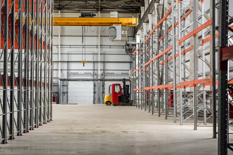 Overhead crane and compact forklift trucks in a empty industrial warehouse building with rows of racks