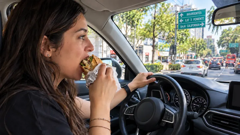 Mujer conduce un automóvil en una avenida de la Ciudad de México mientras da una mordida a una torta y mantiene una mano sobre el volante.