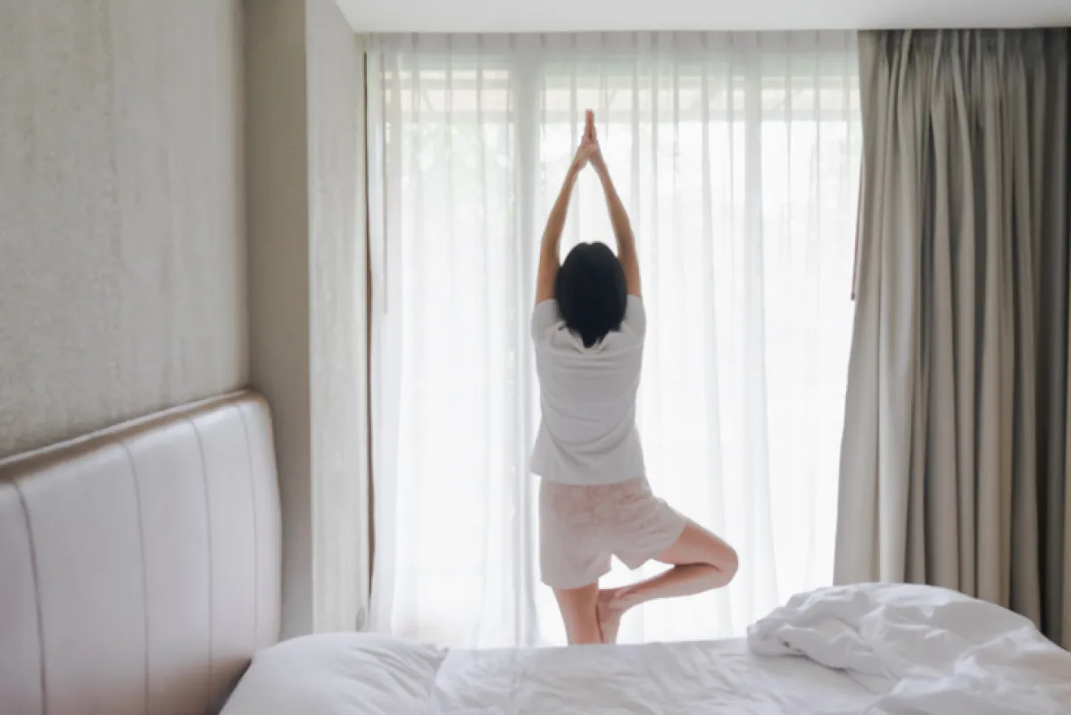 Woman stretching in bed after waking up, back view. Woman sitting near the big white window while stretching on bed after waking up with sunrise at morning, back view.