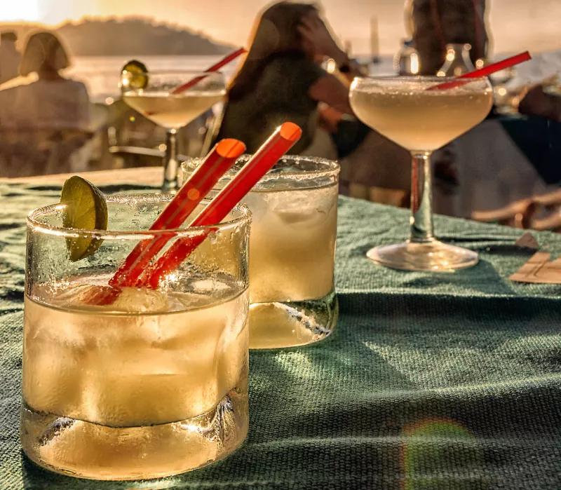 bar de la playa, con cócteles en una mesa, y gente en el fondo viendo la puesta de sol en el océano