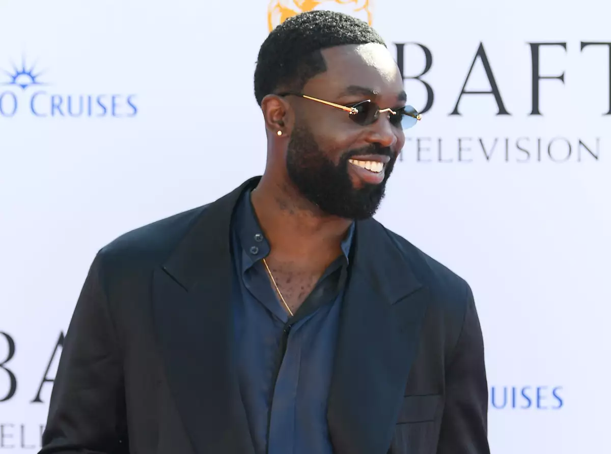 Foto del actor Paapa Essiedu con camisa azul intenso, saco oscuro, barba y lentes de sol, sonriendo en la alfombra roja de los Bafta 2024.