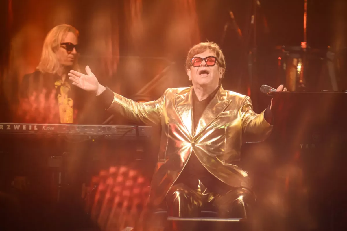 foto de elton john con un saco dorado brillante y los brazos abiertos, sentado junto a su piano.