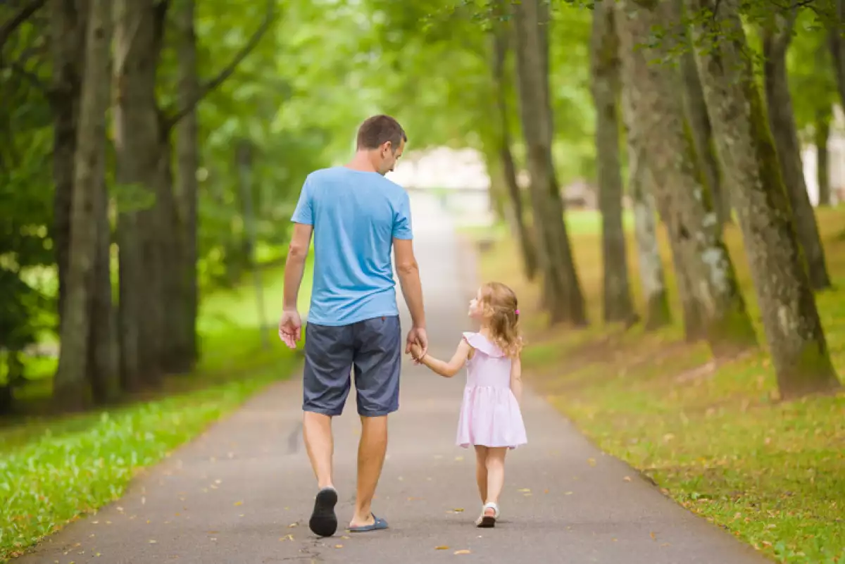 Little daughter and young adult father speaking and walking on sidewalk through tree alley at city park. Spending time together in beautiful warm sunny summer day. Back view.