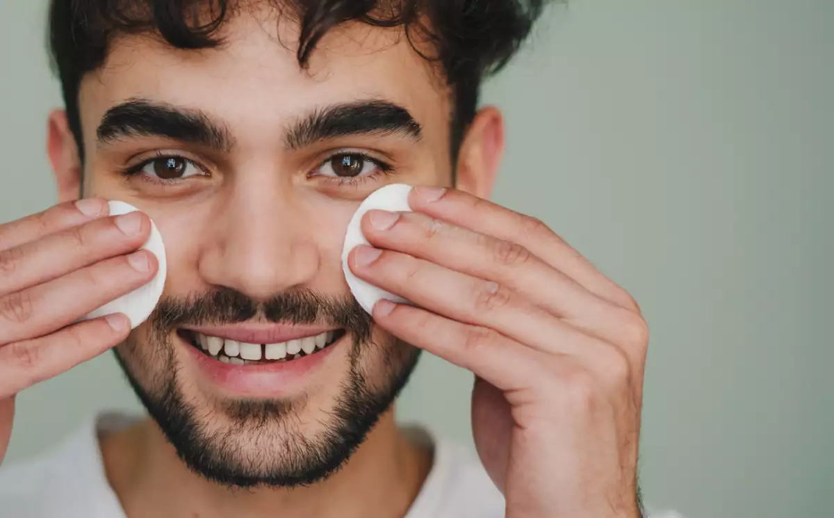 Foto close up del rostro de un hombre joven de barba sonriendo sosteniendo sobre sus mejillas dos algodones de limpieza facial.