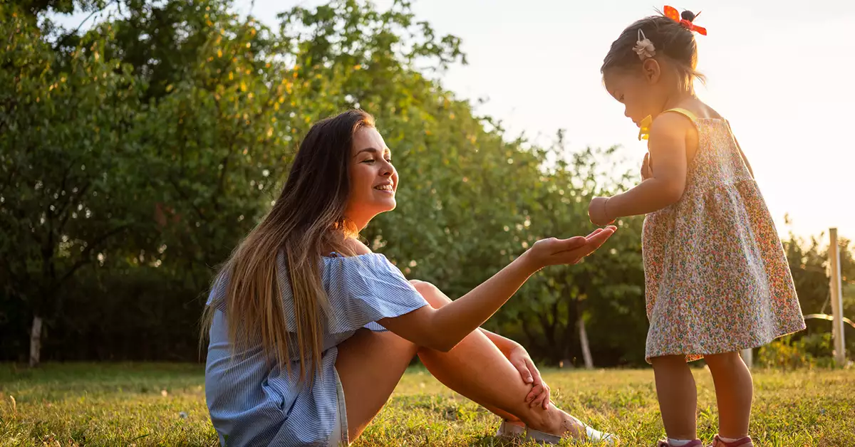 Mujer joven, con vestido azul, sentada sobre el pasto, estirando ligerament brazo derecho, a niña pequeña, con vestido en todo gris blanco