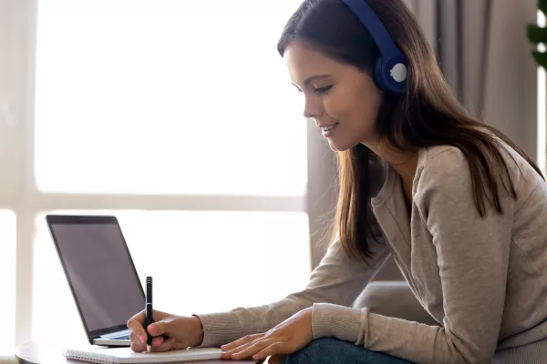 Focused woman in headphones writing notes, using laptop at home