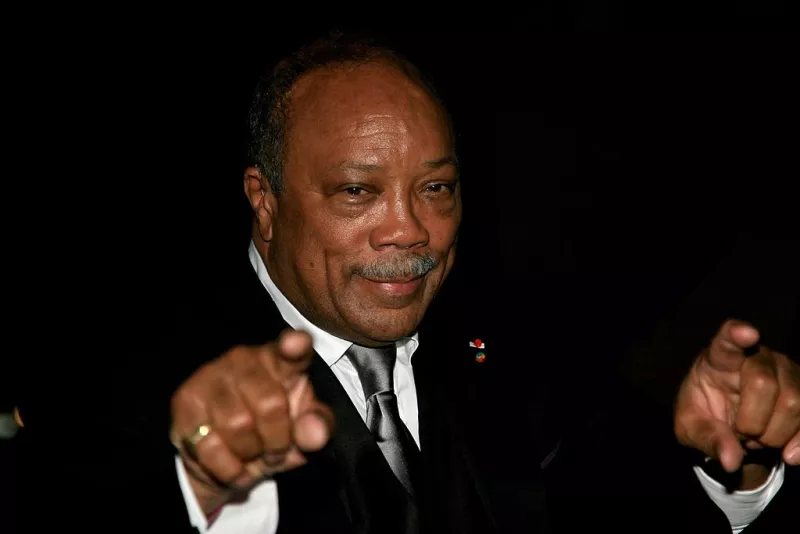 Foto de Quincy Jones en 2007, con traje negro y corbata plateada, sonriendo y apuntando con los dedos índices a la cámara.