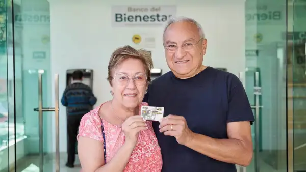 Una mujer y un hombre, ambos adultos mayores, abrazados de lado y sosteniendo una tarjeta del banco Bienestar. 