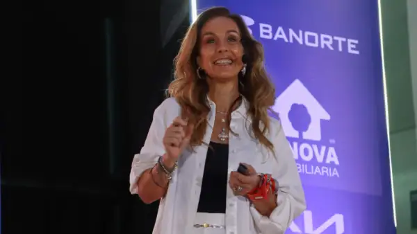 Carla Juan Chelala viste traje blanco y blusa negra. Sonríe, porta un micrófono alámbrico, detrás hay un cártel de Banorte en morado .jpg