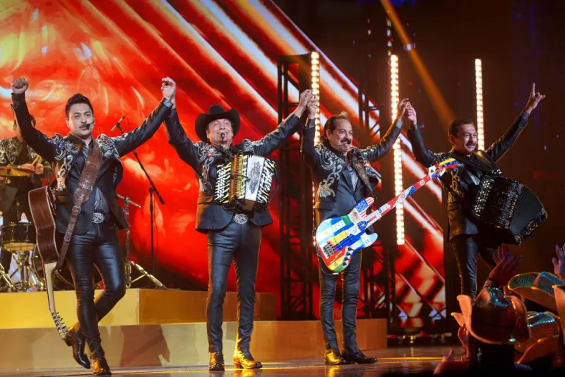 Foto de Los Tigres del Norte en el escenario, con sus instrumentos y tomados de la mano con los brazos en alto.