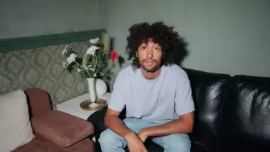 Foto del DJ &Me, sentado en una sala de piel negra, en una habitación de paredes verdes, él viste playera gris y jeans.