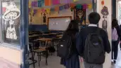 Cuatro jóvenes estudiantes con mochila al hombro saliendo de un salón de clases adornado con papel picado y catrinas.jpg