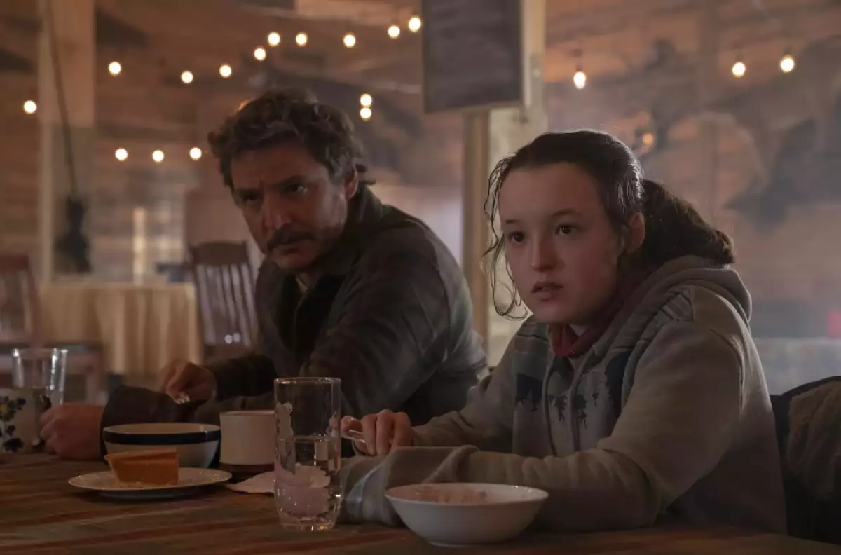 Foto de una escena de la temporada 1 de The Last Of Us con Joel (Pedro Pascal) y Ellie (Bella Ramsey) sentados comiendo.