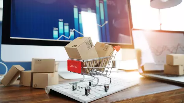 Cajas de papel en carrito de compras  sobre un teclado de computadora, al fondo un gráfico de crecimiento en la pantalla de la computadora.
