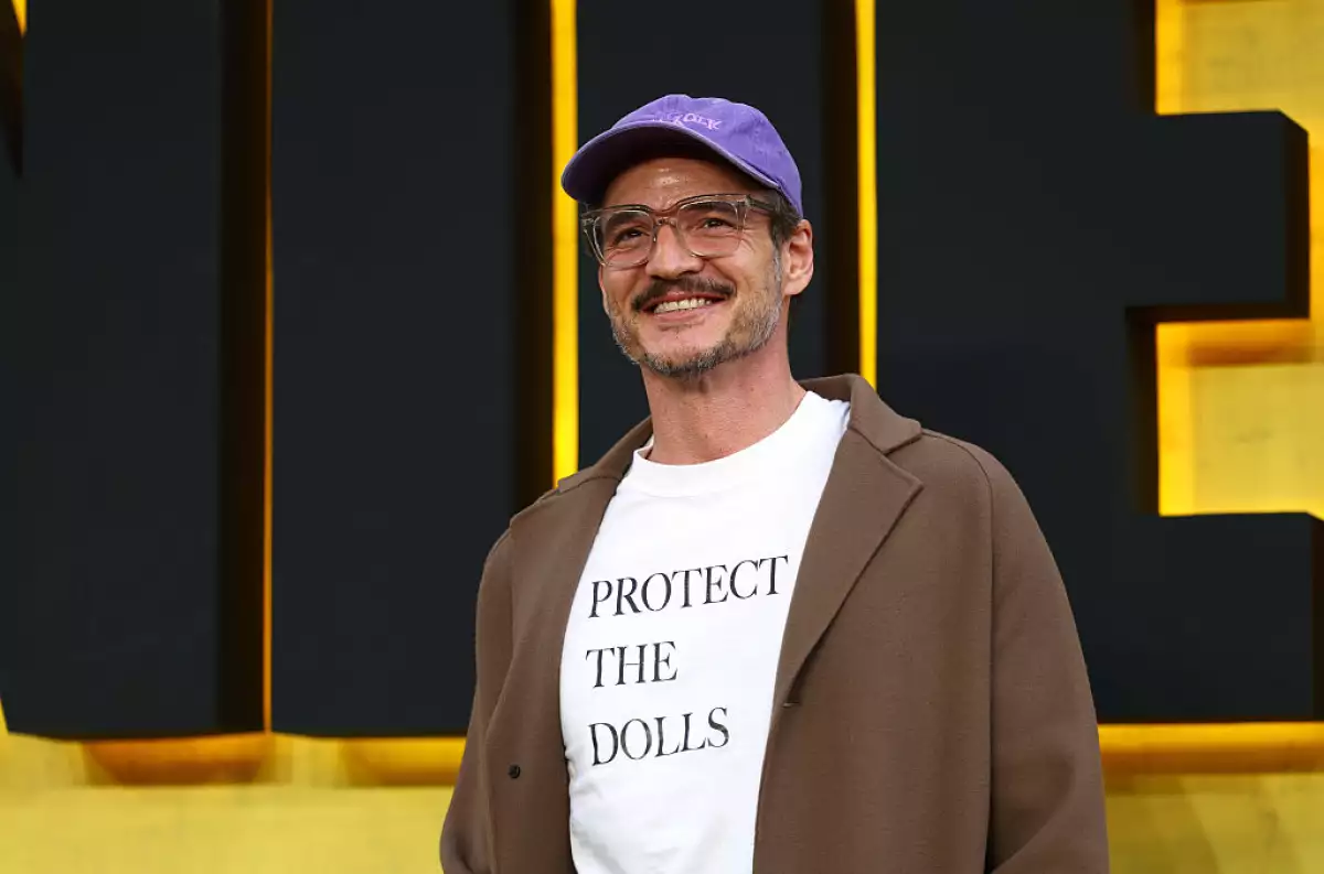 Foto de Pedro Pascal con gorra morada, chaqueta marrón y la playera blanca con letras negras que lleva la leyenda 'Protect the dolls'