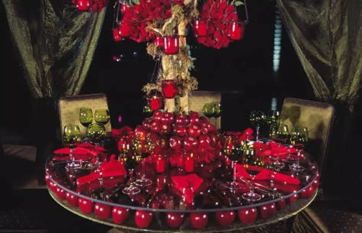 Mesa decorada con arreglo floral de rosas rojas, manzanas y velas del mismo color y copas color verde.