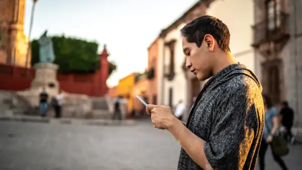 Un joven con con un teléfono en la mano enviando enviando un mensaje en medio de una plaza de un centro histórico. 
