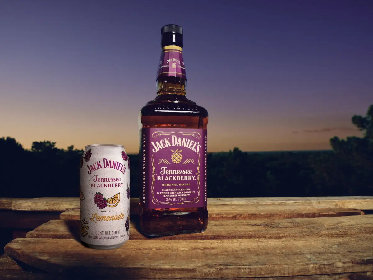 Botella de Jack Daniel’s Tennessee Blackberry y lata de Jack Daniel’s Blackberry Lemonade sobre una superficie de madera, con un paisaje arbolado y el cielo al atardecer como fondo.
