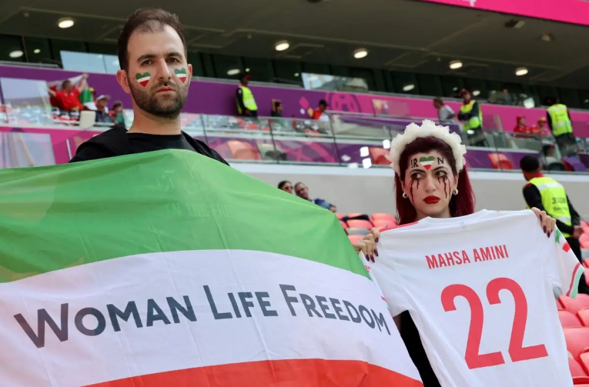 Una mujer con lágrimas maquilladas sostiene una camiseta donde se leer el nombre Mahsa Amini y el número 22, junto con un hombre con corazones pintados en las mejillas sostiene una bandera de franjas horizontales verde, blanca y roja con el mensaje "mujeres, vida, libertad" en las gradas de un partido de Irán en Qatar 2022. 