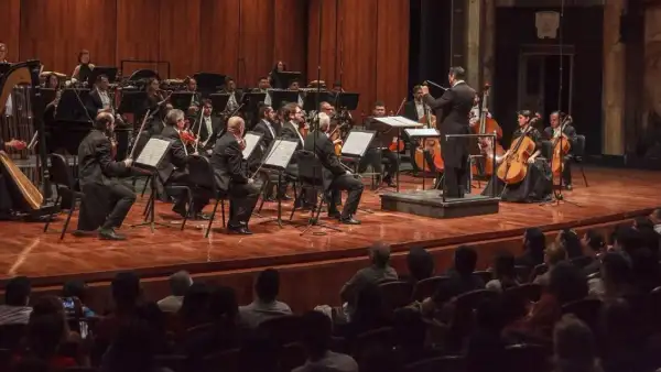 Músicos con violines y partituras con un director de orquesta en un teatro de color café frente a personas sentadasjpg