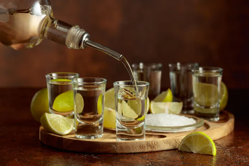 Seis caballitos de tequila sobre una plataforma circular de madera. Una mano sirve tequila blanco. En la tabla redonda también hay limones y sal. 