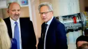 Jensen Huang aparece vestido con un traje azul marino y corbata con puntos durante una reunión. Detrás de él, un aparato tecnológico.