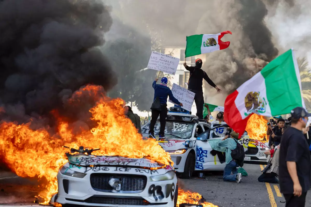 Los manifestantes ondean banderas desde la parte superior de un automóvil destrozado, de pie junto a los vehículos Waymo en llamas, mientras los manifestantes chocan con las fuerzas del orden en las calles que rodean el edificio federal durante una protesta después de las operaciones federales de inmigración en Los Ángeles, California, el 8 de junio de 2025.