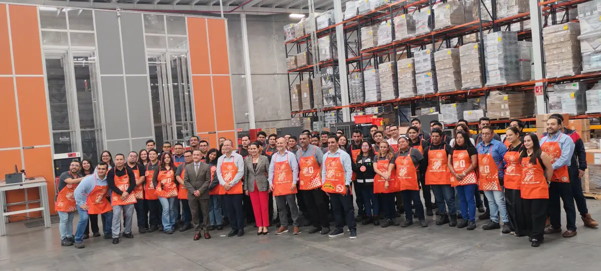 Personal de Home Depot vistiendo los mandiles de dependientes de la cadena en color naranja con el logo de la cadena en color blanco. Están en uno de los centros de distribución de la empresa por lo que hay algunos racks en la parte del fondo. 