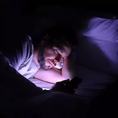 Foto de stock de un hombre acostado en su cama. en un cuarto completamente oscuro y únicamente ilumina su cara el celular