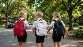 Tres niños rubios de espaldas caminando hacia la escuela por una calle arbolada, vestidos con camisa blanca y pantaloncillos negros.