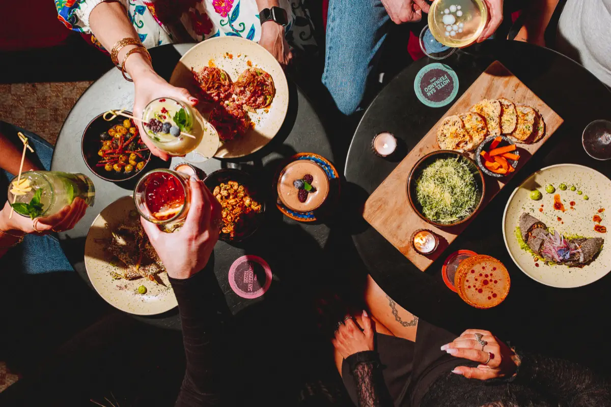 Foto de una mesa con platillos de comida y varias personas con vasos de cocteles en la mano celebrando