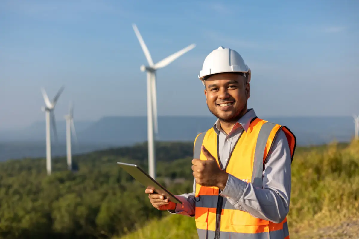 Engineer India man working with tablet at windmill farm Generating electricity clean energy. Wind turbine farm generator by alternative green energy. Asian engineer checking control electric power