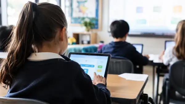 Niños de espaldas utilizan tabletas electrónicas en un salón de clases mientras realizan actividades escolares.