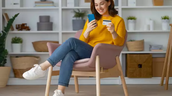 Una mujer sentada en un sillón sostiene su celular  y su tarjeta para compra por WhatsApp Business en el Hot Sale 