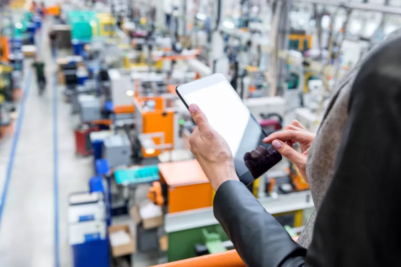 Imagen horizontal en color de una mujer de negocios, una persona irreconocible, trabajando con una tableta digital en una gran fábrica futurista. Mujer de pie en la parte superior de un balcón, sosteniendo un panel táctil y comprobando el inventario de una empresa de fabricación en una tableta con pantalla táctil. Concéntrate en las manos de la mujer de negocios sosteniendo una tableta negra, máquinas futuristas con una persona incidental en el fondo.