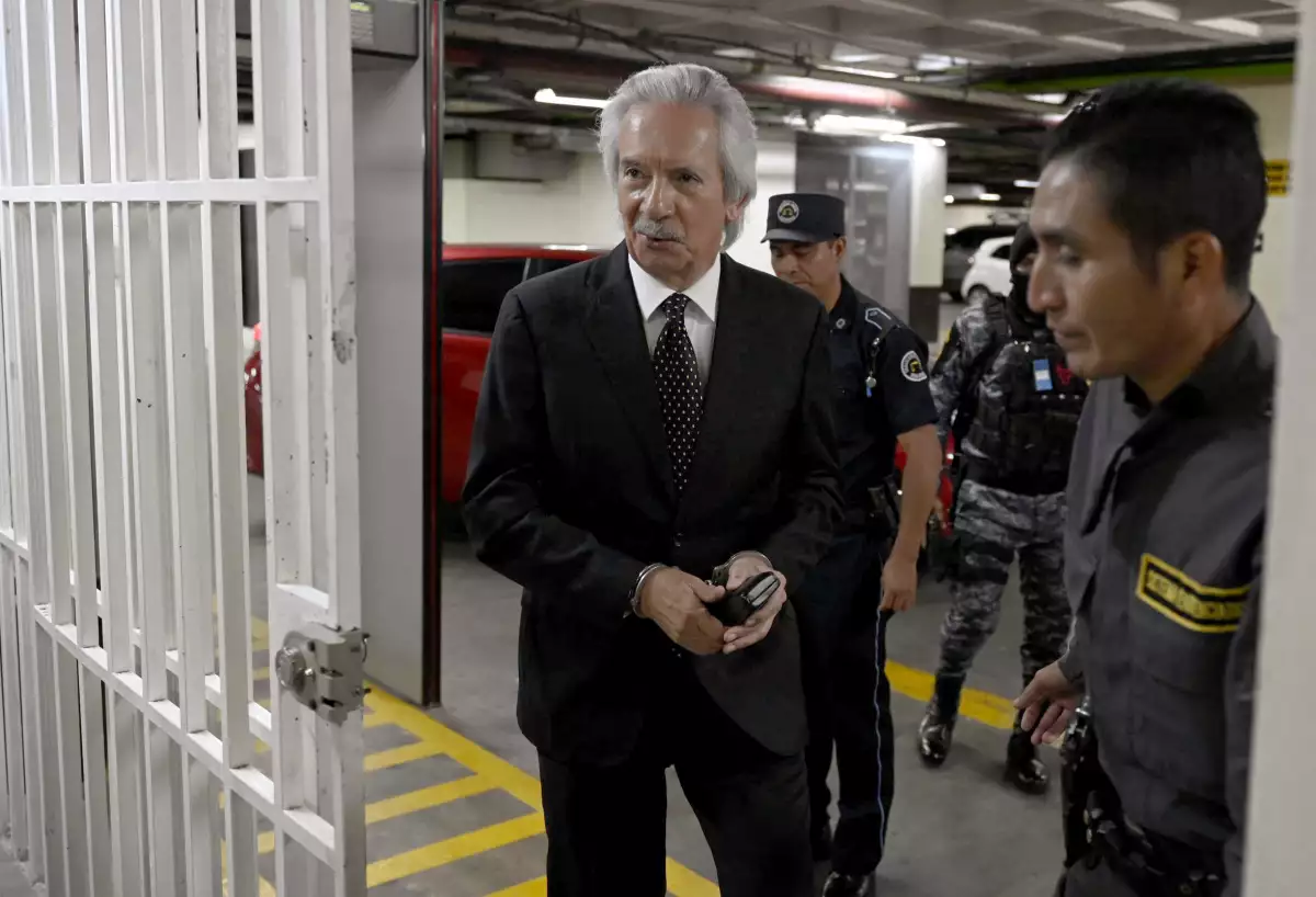 El periodista guatemalteco, José Ruben Zamora de 66 años. Lleva un traje negro, camisa blanca y corbata con puntos, se encuentra esposado. 
Lo escoltan al menos 3 hombres parte de la fuerza militar y policiaca.