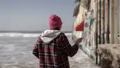 Una persona sostiene una bandera de México con su mano derecha en el muro fronterizo entre México y Estados Unidos.