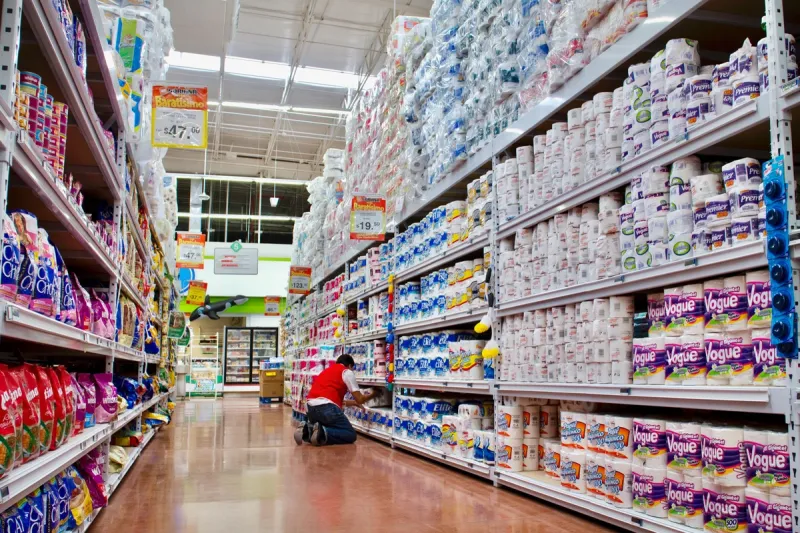Interior de un supermercado con un amplio pasillo dedicado a productos de papel y artículos de limpieza. A ambos lados del pasillo se observan estanterías metálicas altas, completamente abastecidas con paquetes de papel higiénico, servilletas, toallas de papel y productos de limpieza de distintas marcas. En el lado derecho predominan grandes cantidades de rollos y paquetes de papel higiénico, mientras que en el lado izquierdo se exhiben productos de limpieza y paquetes de artículos para el hogar. En el centro del pasillo, un trabajador del supermercado, vestido con pantalón de mezclilla, camiseta blanca y chaleco rojo, se encuentra agachado frente a los estantes mientras acomoda mercancía. 
