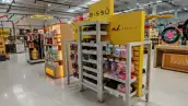 nterior de una tienda OfficeMax con un exhibidor amarillo de Bissú y productos de maquillaje y belleza distribuidos en estantes. Al fondo se observan otras áreas de venta y pasillos de la sucursal. La imagen muestra la presencia de categorías de cuidado personal dentro del establecimiento.