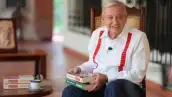 Andrés Manuel López Obrador sostiene dos libros en sus manos, viste guayabera blanca con vivos rojos y pantalón azul.jpg