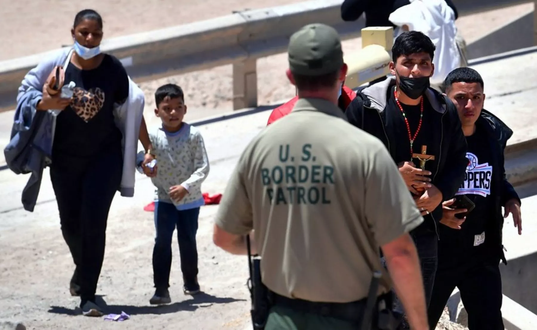 (Un migrante sostiene su crucifijo al cruzar un puente hacia el muro fronterizo, mientras un oficial de Aduanas y Protección Fronteriza (CPB) de EE. UU lo observa)