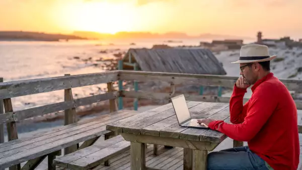 Nómada digital sentado al aire libre en la playa con un portátil solo haciendo teletrabajo al atardecer